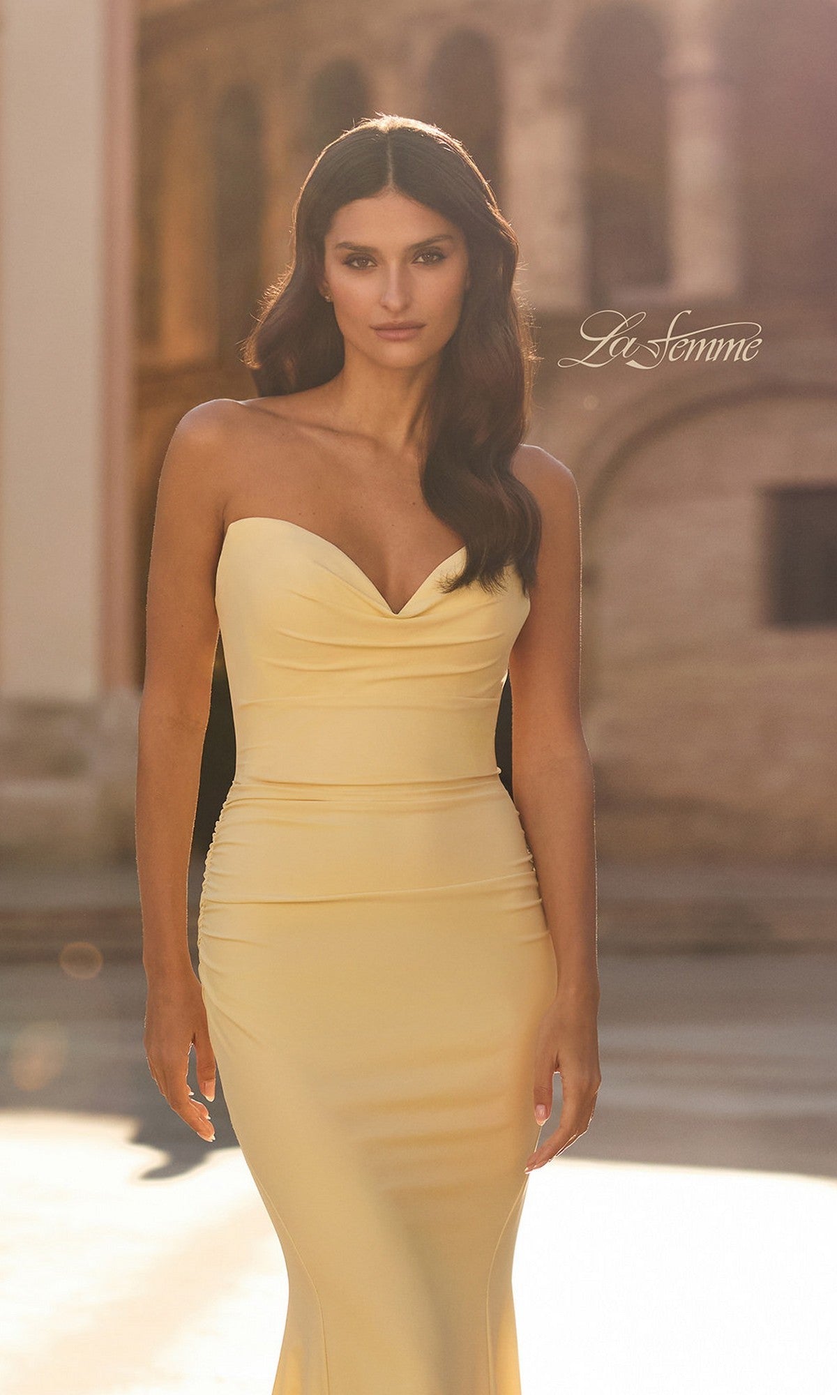 A woman stands outdoors in natural light, wearing the yellow La Femme 33864 long strapless jersey prom dress with a softly blurred historic building in the background. The La Femme logo appears in the image.